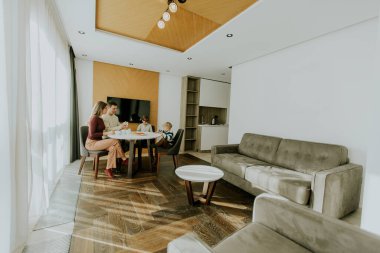 Parlak bir oturma odasında bir aile bir masanın etrafında oturur. Kahvaltıyı paylaşıyorlar ve konuşuyorlar. Oda açık renkli ve yumuşak mobilyalı modern bir tasarıma sahip. Pencereden güneş ışığı geliyor..