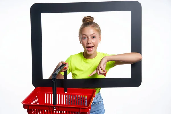オンラインショッピングのコンセプト 流行のティーン女の子を通して見てデジタル画面を保持空のショッピングバスケット 驚きとそれを指して 光の背景に隔離 — ストック写真