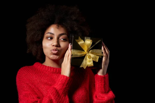黑色星期五销售 节日礼物 庆祝概念 一个快乐的非洲裔美国女人拿着一个带着金色蝴蝶结的黑色礼品盒 在黑色背景下亲吻着它 — 图库照片