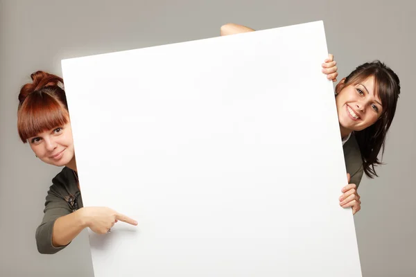 Two young women peeking over edge of blank empty paper billboard — Stok fotoğraf