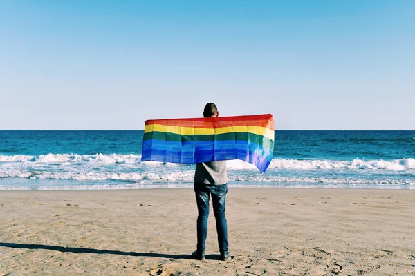 Hombre con una bandera de arco iris frente al océano — Foto de Stock