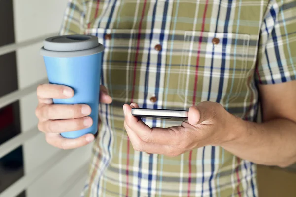 Человек с чашкой кофе или чая с помощью смартфона — стоковое фото