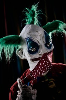 Yeşil saçlı, kirli kırmızı bir kostüm, kirli bir papyon ve kanlı eldivenler giyen, sirk çadırının önünde kırmızı bir yüz maskesi takan ya da çıkaran rahatsız edici kötü bir palyaçonun yakın çekimi.