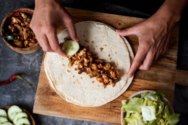 Kafkasyalı genç bir adamın soğan, kırmızı ve yeşil biber, taze marul ve çiğ salatalık gibi farklı sebzelerle pişirilmiş tavuk eti ya da burrito hazırlarken yüksek açılı görüntüsü.