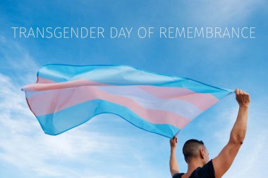 closeup of a young caucasian person, seen from behind, holding a transgender pride flag on the air and the text transgender day of remembrance on the sky clipart
