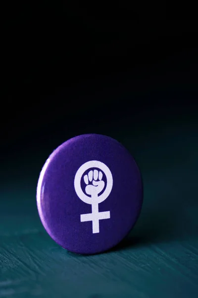 the women power symbol, a raised fist in a female gender symbol, in a violet pin button on a dark gray surface, against a black background with some blank space on top