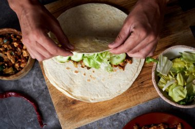 Kafkasyalı bir adamın, soğan, kırmızı ve yeşil biber, taze marul ve çiğ salatalık gibi farklı sebzelerle pişirilmiş tavuk eti ya da burrito yuvarlarken yüksek açılı görüntüsü.