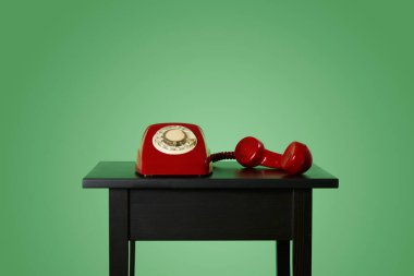 Kırmızı sabit hatlı, yeşil arka planda, ahşap bir masada, ahşap ahizeden kopmuş bir cep telefonu.
