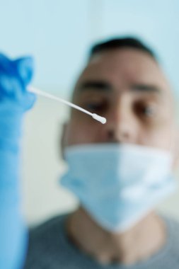 Mavi ameliyat eldiveni takan bir sağlık görevlisinin, ağzını kapatan bir maske takmış, genç bir adamın burnundan burun sıvısı almak üzere olan bir fotoğrafı.