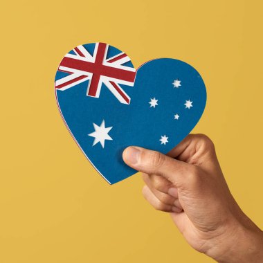Elinde kalp şeklinde bir Avustralya bayrağı tutan bir adamın eli. Sarı arka planda, farklı renkte kağıt kesikleriyle yapılmış.