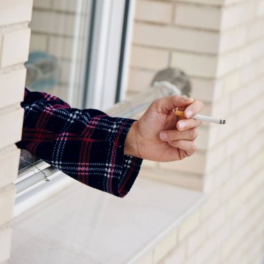 Sıcak pijama giymiş, evinin veya otel odasının camında sigara içen genç bir beyaz adamın yakın çekimi.