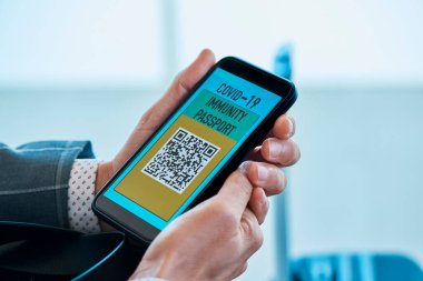 Zarif gri takım elbiseli genç beyaz bir işadamı, havaalanının bekleme salonunda oturuyor, akıllı telefonunun ekranında simüle edilmiş elektronik covid-19 dokunulmazlık pasaportu var.