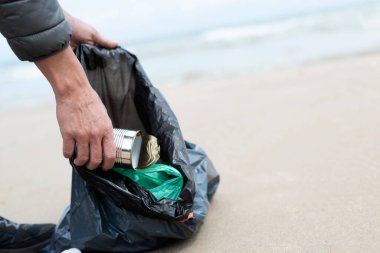 Genç bir beyaz adam, doğal çevreyi temizlemek için kumsalın kumlarından teneke, şişe ya da plastik torbalar gibi bazı atıkları topluyor.