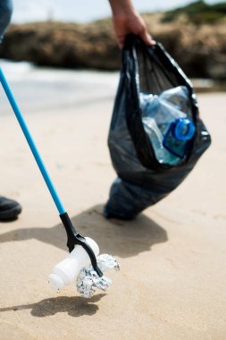 Bir adam, doğal çevreyi temizlemek için kumsalın kumlarından teneke, şişe ya da torba gibi daha geniş bir alanda atık toplar.