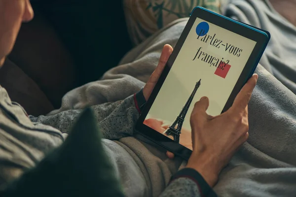 暖かいグレーの毛布に包まれたソファに横になってタブレットを見ている若い白人男性が閉じ込められていますフランス語で書かれたフランス語を読みますか — ストック写真