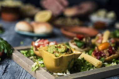 Atıştırmalık ya da aperatif olarak sunulan farklı malzemelerle vejetaryen sandviçlerin yanında guacamole olan bir kaseye yakın plan gri bir masada ve arka planda yemek hazırlayan bir adam.