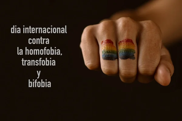 在年轻人的拳头上画的彩虹旗和在黑色背景下以西班牙语书写的反对恐同 变性和恐惧症国际日的文字 — 图库照片