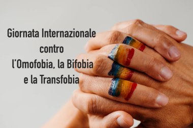 Genç bir insanın sıkışık ellerine çizilmiş gökkuşağı bayrağı ve beyaz bir arkaplanda İtalyanca yazılmış homofobi, transfobi ve bifobiye karşı uluslararası yazı.