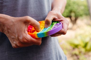 closeup of a young caucasian man, wearing a casual grayish t-shirt outdoors, popping some bubbles of a destressing toy patterned with the colors of the rainbow clipart