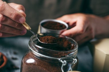 Kafkasyalı genç bir adamın, gri bir ahşap masada, cam kavanozdan yapılmış kahveyle bir çaydanlığın hunisini doldurması.