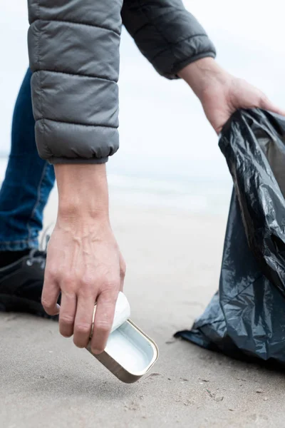一个拿着垃圾袋的年轻人用手从海滩的沙地里捡起一只用过的食物罐 作为对自然环境的一种净化行动 — 图库照片