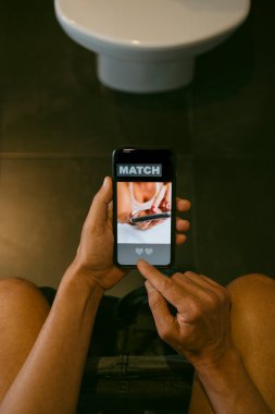 Kafkasyalı bir adamın yüksek açılı görüntüsü, tuvalette oturuyor, akıllı telefonuyla internetten flört ediyor, ekranında kelime eşleşmesi olan sahte bir uygulama var.