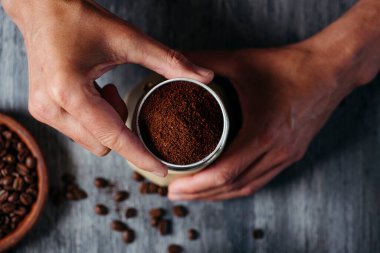 Kafkasyalı genç bir adamın moka demliğine kahve dolu huniyi yerleştirirken yüksek açılı görüntüsü, gri bir ahşap masaya yerleştirilmiş.