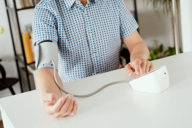 Beyaz bir adam kan basıncını elektronik bir sfenometre ile ölçüyor. Evdeki beyaz bir masada oturuyor.