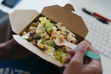 Kafkasyalı genç bir adam, masasında bambu çatalı kullanarak kağıt bir kabın içinden nohut salatası yiyor.