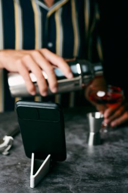 Kafkasyalı genç bir adamın yakın çekimi, çizgili bir gömlek giyiyor, metal bir karıştırıcıda kokteyl hazırlıyor, akıllı telefonundaki özel bir dersi takip ediyor.