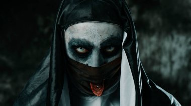 Siyah beyaz bir giysi giyen korkunç bir rahibeye yakın çekim, siyah yüz maskesindeki delikten dilini çıkarıyor.