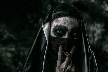 Tipik siyah beyaz giysili korkunç bir rahibe, siyah maskesindeki delikten dilini çıkarır.