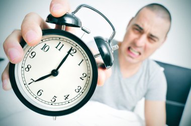 angry young man in bed holds his alarm clock clipart