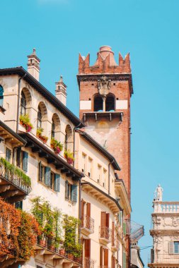Torre del Gardello kulesi açık mavi bir gökyüzünün altında güneşli bir günde İtalya 'nın Verona şehrinde balkonları ve yeşil kepenkleri olan renkli tarihi binaların üzerinde yükseliyor.