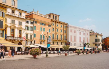 Verona, İtalya - 24 Haziran 2025: Renkli binalar Piazza Bra 'nın önünden geçiyor, insanlar sıcak güneşin ve merkez Verona' nın canlı atmosferinin tadını çıkaran lokantalarla dolu canlı kafe teraslarının önünden geçiyor.