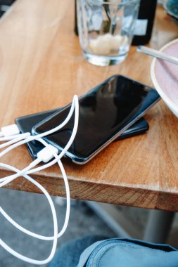 Tahta bir masanın üzerinde uzanan akıllı telefonun yakın görüntüsü. Elektrik bankasına dolanmış bir kablo kullanılmış bulaşıklar ve açık hava yemeğinden sonra bir bardak ile çevrili.