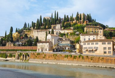 Verona, İtalya - 23 Haziran 2025: Adige nehri boyunca yürüyen ziyaretçiler, üzerlerindeki yamaç ve tarihi evlere tepeden bakan selvi ağaçları ile çevrili olan Castel San Pietro.