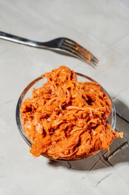 Yuvarlak cam bir kâse dolusu parçalanmış portakal soslu domuz eti, desenli gri bir yüzeye yerleştirilirken çatal yumuşak bir gölgenin üzerinde duruyor.