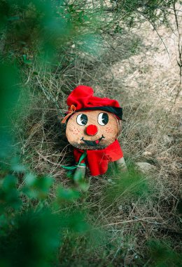 İspanya 'daki Katalonya' dan küçük bir Tio de Nadal karakteri, kısmen ağaçlık alanda gizlenmiş kırmızı bir şapkası, boyalı bir gülümsemesi ve şenlikli kırmızı bir eşarbı var.