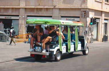 Pisa, İtalya - 26 Haziran 2025: Küçük bir elektrikli tur aracı Lungarno Mediceo boyunca yeşil gölgeliğinin altında oturan bir grup turistle birlikte şehir merkezinin tarihi binalarının önünden geçiyor.
