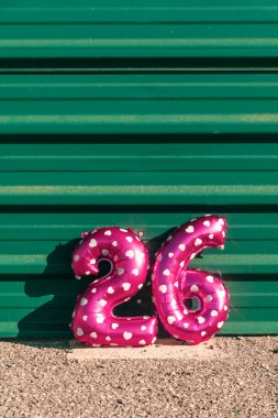 26 numara biçimindeki pembe folyo balonlar yeşil metal bir duvarın üzerinde duruyor. Beyaz kalp desenleri, neşeli bir kutlama sahnesinde gün ışığı altında parlıyor.