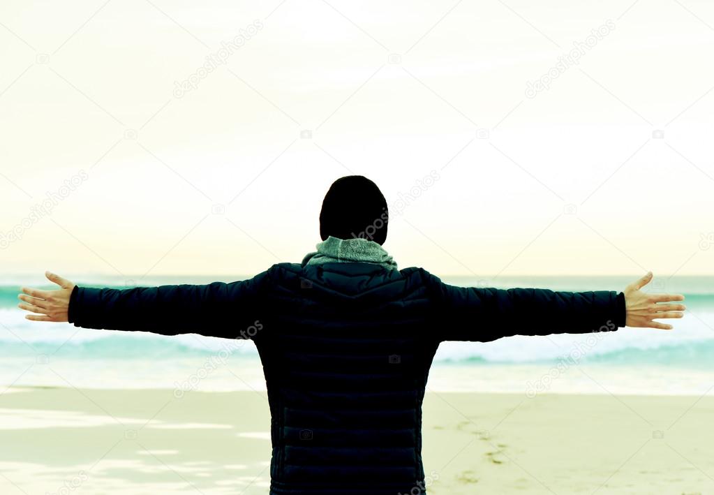 man in coat with his arms in the air in front of the ocean