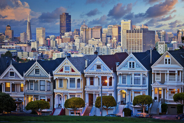 Famous Painted Ladies of San Francisco, California sit glowing amid the backdrop of a sunset and skyscrapers. 