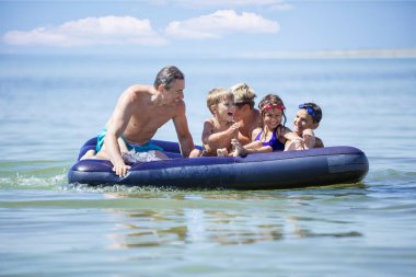 Kardeşler, arkadaşları ve babaları şişme şişme yatakta eğleniyorlar. Küçük çocuklar ve babalar deniz kenarında tatilin tadını çıkarıyorlar..