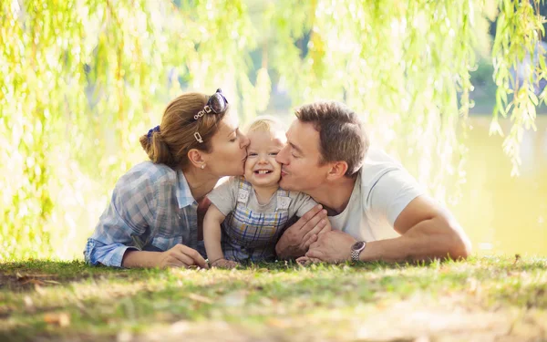 Gelukkige ouders zoenen peuter jongen. Vader, moeder en zoon ontspannen op het gazon. — Stockfoto