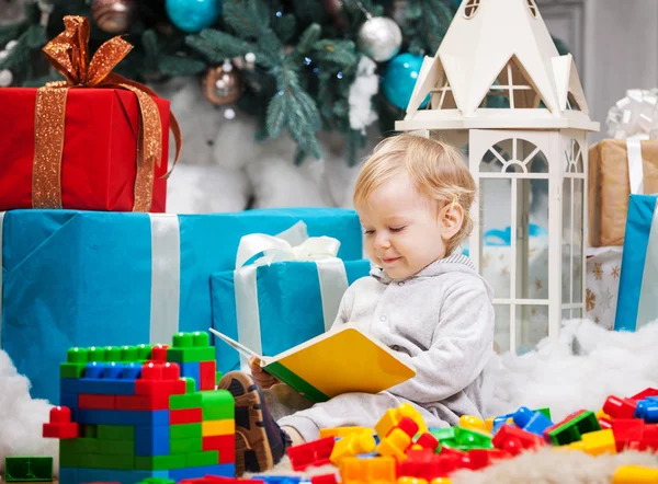 Lindo niño sentado en el árbol de Navidad y leyendo el libro. Bloques de construcción dispersos alrededor — Foto de Stock