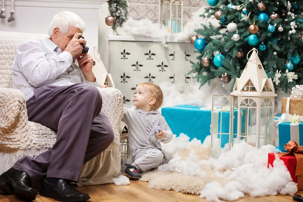 老人拍照的他蹒跚学步的孙子坐在圣诞树在家附近的 — 图库照片