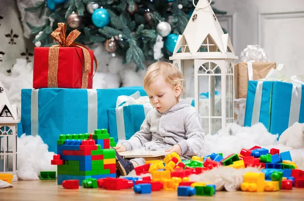 Cute toddler boy sitting at Christmas tree and reading book. Building blocks scattered around. — Stock Photo, Image