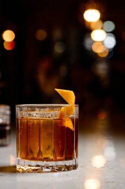 Yakın plan vaftiz baba kokteyli. Parlak bar arka planında bir dilim portakal ve buz var. Alkolik Baba, portakal kabuğu ve buzlu.