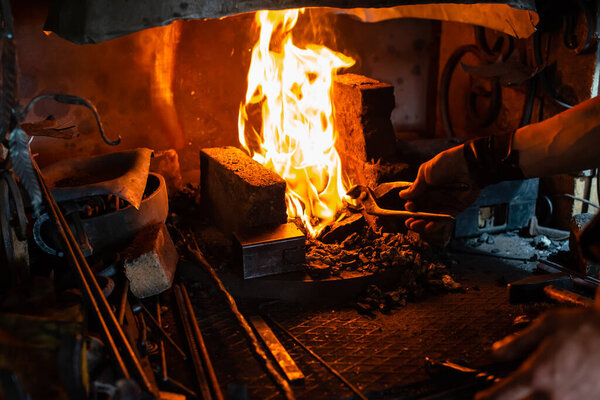 The blacksmith holds billet over hot coals in clay oven. blacksmith heating iron metal sword manufacturing marching forge, hands of a blacksmith hold piece of steel in fire of a red-hot forge
